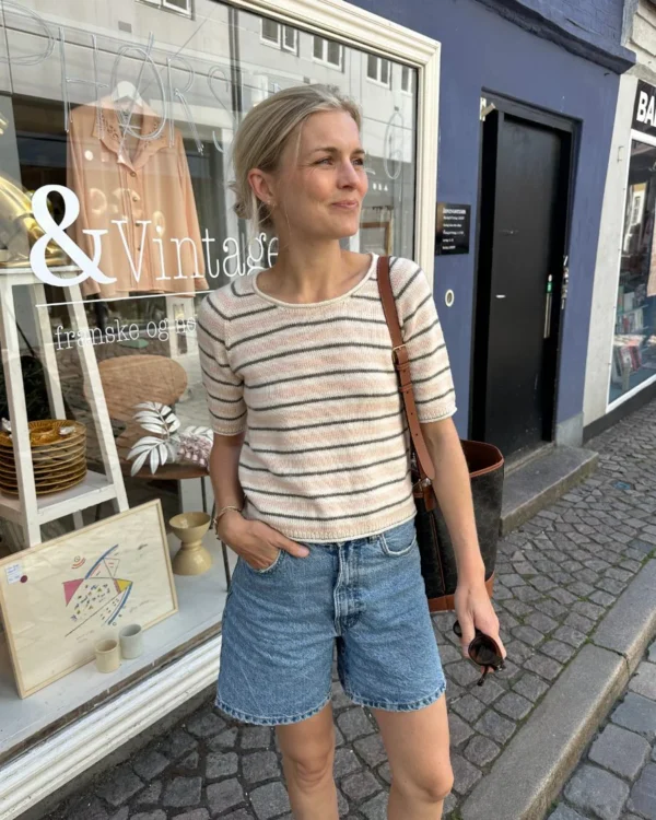 Kledd i en Amélie-t-skjorte og denimshorts står en person på et brosteinsbelagt fortau foran en vintagebutikk.