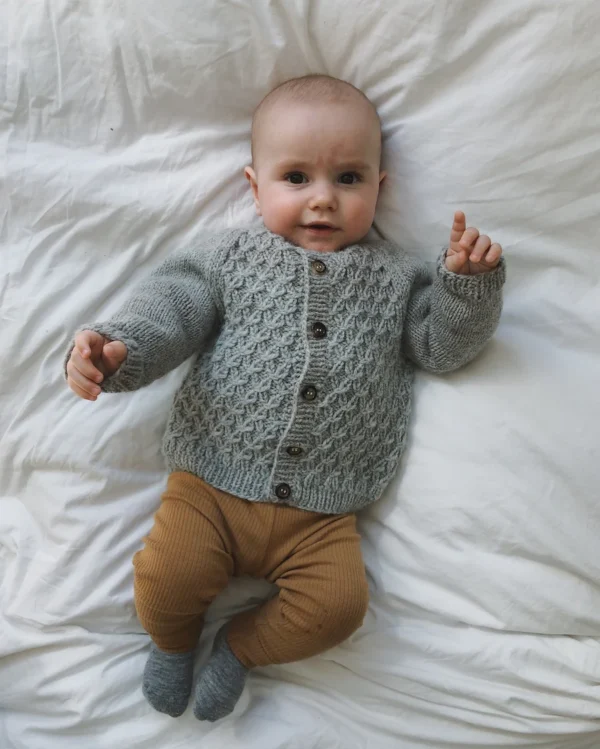 En baby kledd i en tettsittende Carls cardigan og brune bukser ligger på en hvit seng og stirrer oppover med den ene hånden forsiktig hevet.