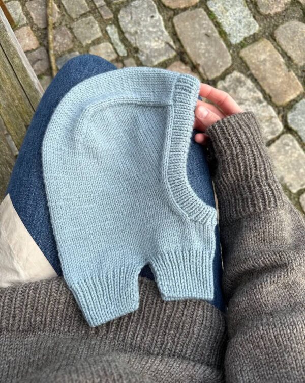 Sittende på en benk holder en person et lyseblått strikket babyplagg mens han har på seg Everyday Balaclava sammen med en grå genser og jeans. En brosteinssti utfolder seg i bakgrunnen.