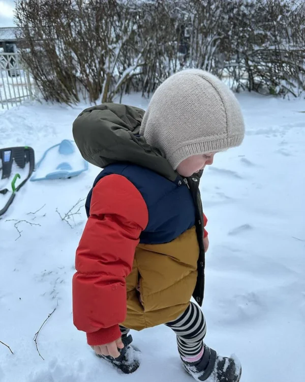 Et barn i en livlig jakke og lue, med en Everyday Balaclava subtilt synlig, går gjennom snøen mens snøskuffene står rett i bakgrunnen.