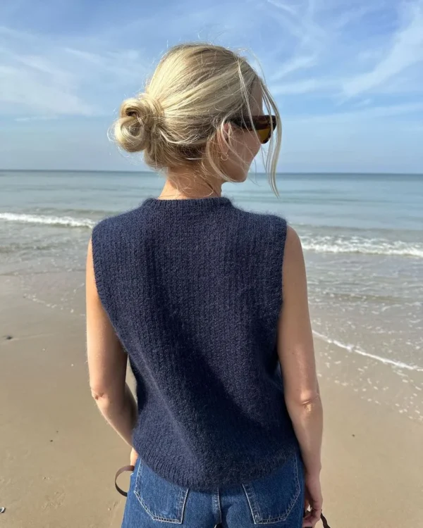 En person med blondt hår i en bolle, sportslige solbriller, den elegante fredagsslipoveren i marineblått og jeans står på stranden med utsikt mot havet. Det er det perfekte tilfluktsstedet for denne stilige strandgjengeren.