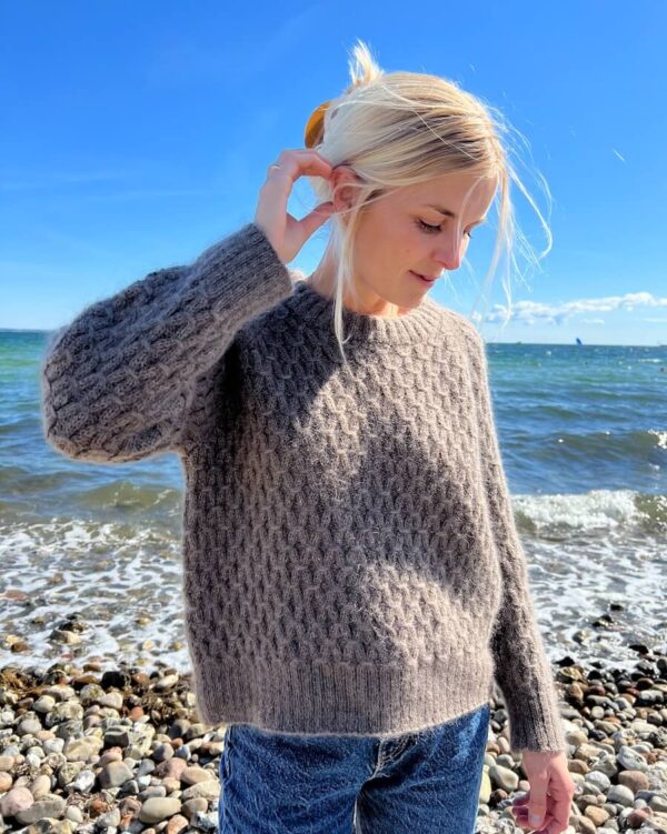 Jenny, hennes blonde hår forsiktig rufset av havbrisen, står på en steinete strand iført en moteriktig Jenny-genser. Den grå strikken kompletterer jeansen hennes, og blander seg vakkert med det store havet og den blå himmelen i bakgrunnen.