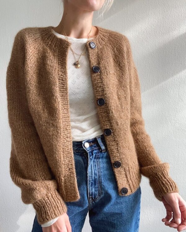 En nybegynner modellerer den brune Novice Cardigan - Chunky Edition, og parer den enkelt sammen med en hvit topp og blå jeans.