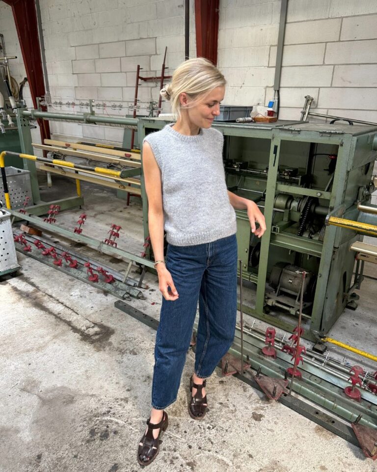 En person iført en nybegynner slipover og jeans står i industrielle omgivelser, med ruvende maskineri i bakgrunnen.