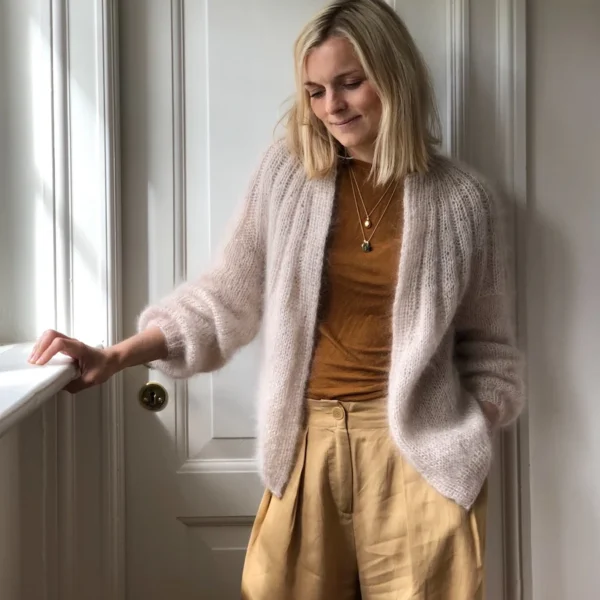 En person med lyst hår, pakket inn i en Sunday Cardigan - Mohair Edition, og iført en brun skjorte står ved en hvit dør og stirrer litt ned.