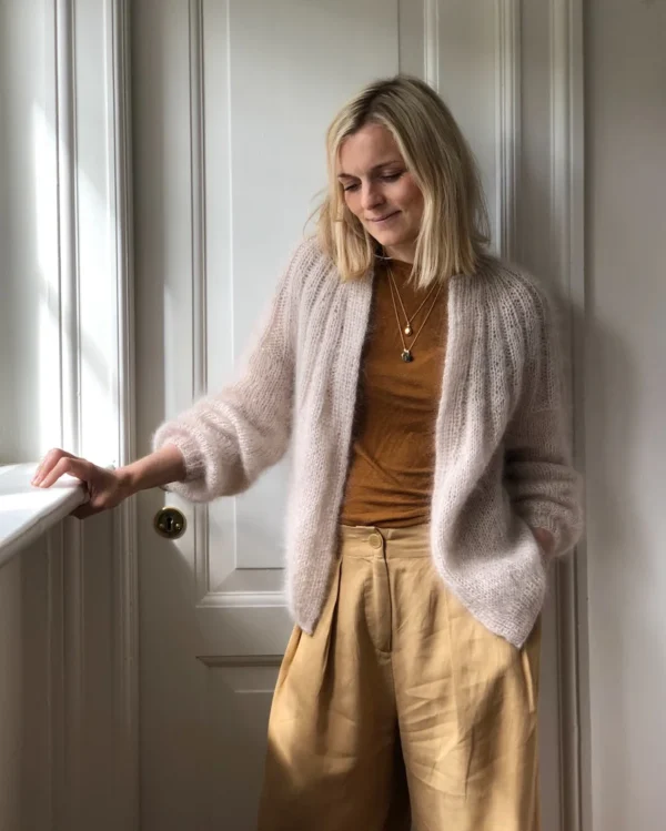 En person med lyst hår, pakket inn i en Sunday Cardigan - Mohair Edition, og iført en brun skjorte står ved en hvit dør og stirrer litt ned.