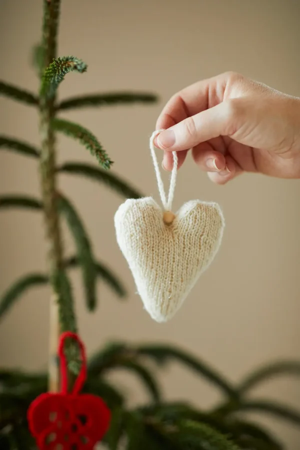 En hånd holder forsiktig et hjerteformet ornament kjent som "454 Jul i norsk ull", laget av norsk ull, nær en juletregren.