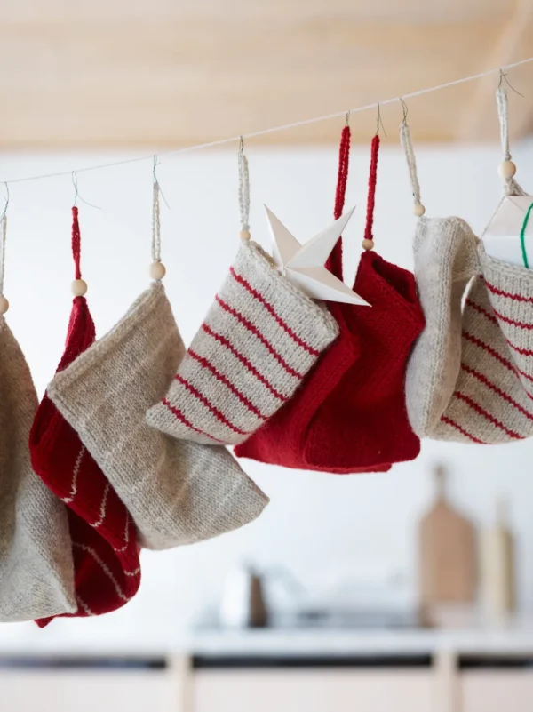 Røde og grå julestrømper, omhyggelig laget av norsk ull, henger på en linje utsmykket med stjernepynt i en mykt fokusert innendørs setting, og tilfører scenen et element av 454 Jul i norsk ull.