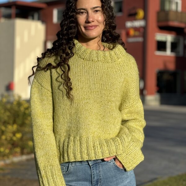 En person iført en lysegrønn genser og jeans står utenfor med en bygning i bakgrunnen.