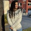 En person med krøllete hår, iført en Hjertegenser voksen XXL-mønstret genser og jeans, står ved et tre og smiler. I bakgrunnen fullfører en rød bygning og parkert bil åstedet.
