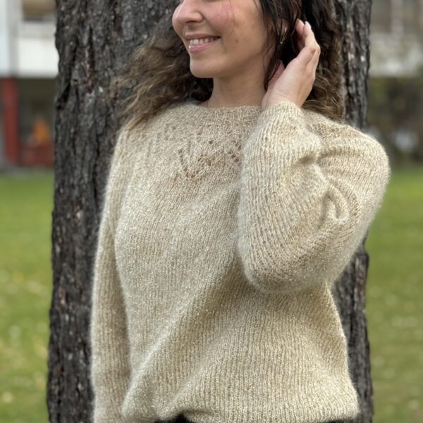 En person med langt hår smiler mens han står nær et tre, iført en beige genser og svarte bukser.