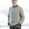 En person iført en mønstret genser og jeans fra Produktnavn: 2305 står innendørs og smiler med snødekt vindusbakgrunn.