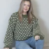 En person sitter på en sofa, iført en grønn- og hvitmønstret genser og jeans, med langt hår som faller ned. Deres nøytrale uttrykk forsterker den tidløse stemningen i scenen, og fremkaller en kunstnerisk skildring fra 2305-kolleksjonen.