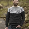 En mann med skjegg står utendørs, komfortabelt kledd i en strikket genser med et svart-hvitt mønster, sammen med blå jeans, og fanger perfekt essensen av "Flokken"-temaet for hele familien.