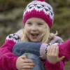 Et barn fra «Flokken»-kolleksjonen smiler varmt, iført rosa strikkelue og genser prydet med hvitt og svart mønster, mens de sitter utendørs blant gress og steiner.