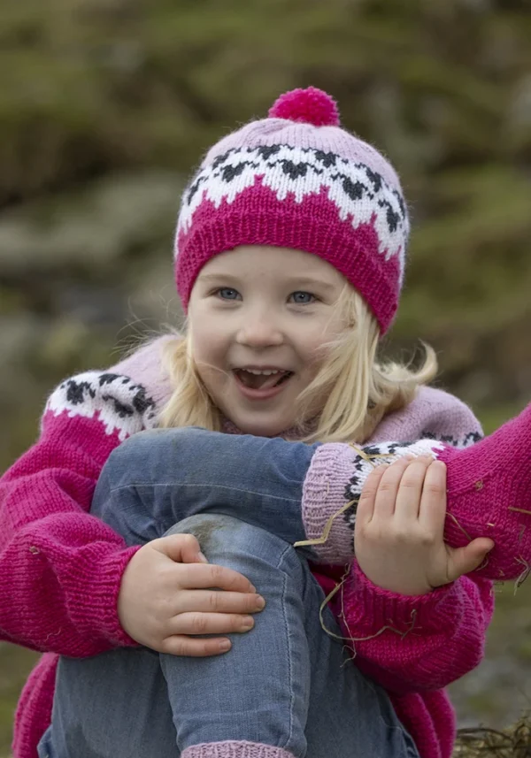 Et barn fra «Flokken»-kolleksjonen smiler varmt, iført rosa strikkelue og genser prydet med hvitt og svart mønster, mens de sitter utendørs blant gress og steiner.