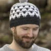 En mann, sannsynligvis en del av "Flokken"-kolleksjonen for hele familien, har på seg en mønstret strikkelue med pompom. Han smiler varmt med lukkede øyne, pakket tett inn i genseren.