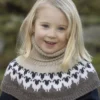 Et lite barn med blondt hår har på seg en beige og svart mønstret strikkegenser, smilende utendørs, som en del av 2309 Tema: "Flokken" til hele familien nyter en gledelig dag i naturen.