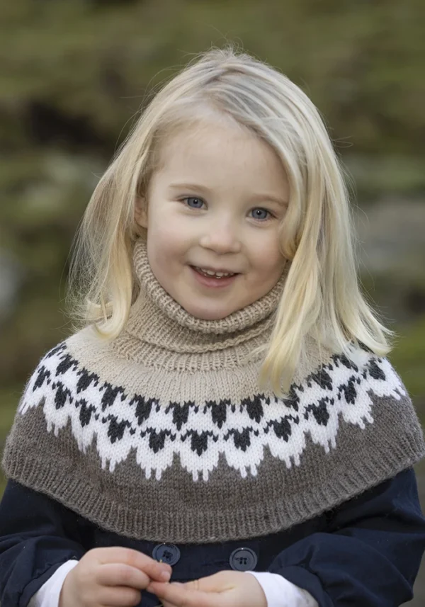 Et lite barn med blondt hår har på seg en beige og svart mønstret strikkegenser, smilende utendørs, som en del av 2309 Tema: "Flokken" til hele familien nyter en gledelig dag i naturen.