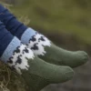Et nærbilde av en iført grønne strikkede sokker prydet med svart-hvitt mønster, sittende på en høyballe, legemliggjør det koselige temaet "Flokken" til hele familien.