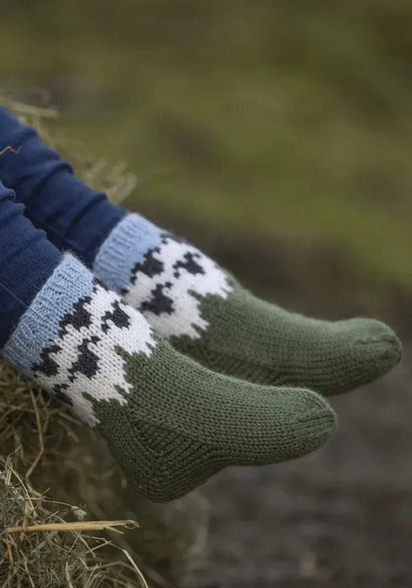 Et nærbilde av en iført grønne strikkede sokker prydet med svart-hvitt mønster, sittende på en høyballe, legemliggjør det koselige temaet "Flokken" til hele familien.