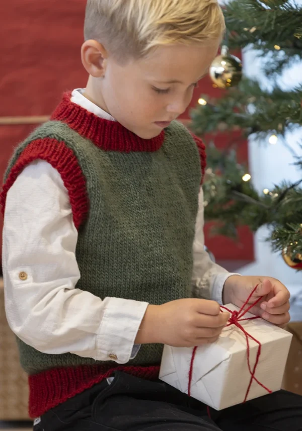 En ung gutt i en festlig genser åpner ivrig en innpakket gave ved siden av et vakkert dekorert juletre, og legemliggjør den gledelige ånden til Tema: Jul.