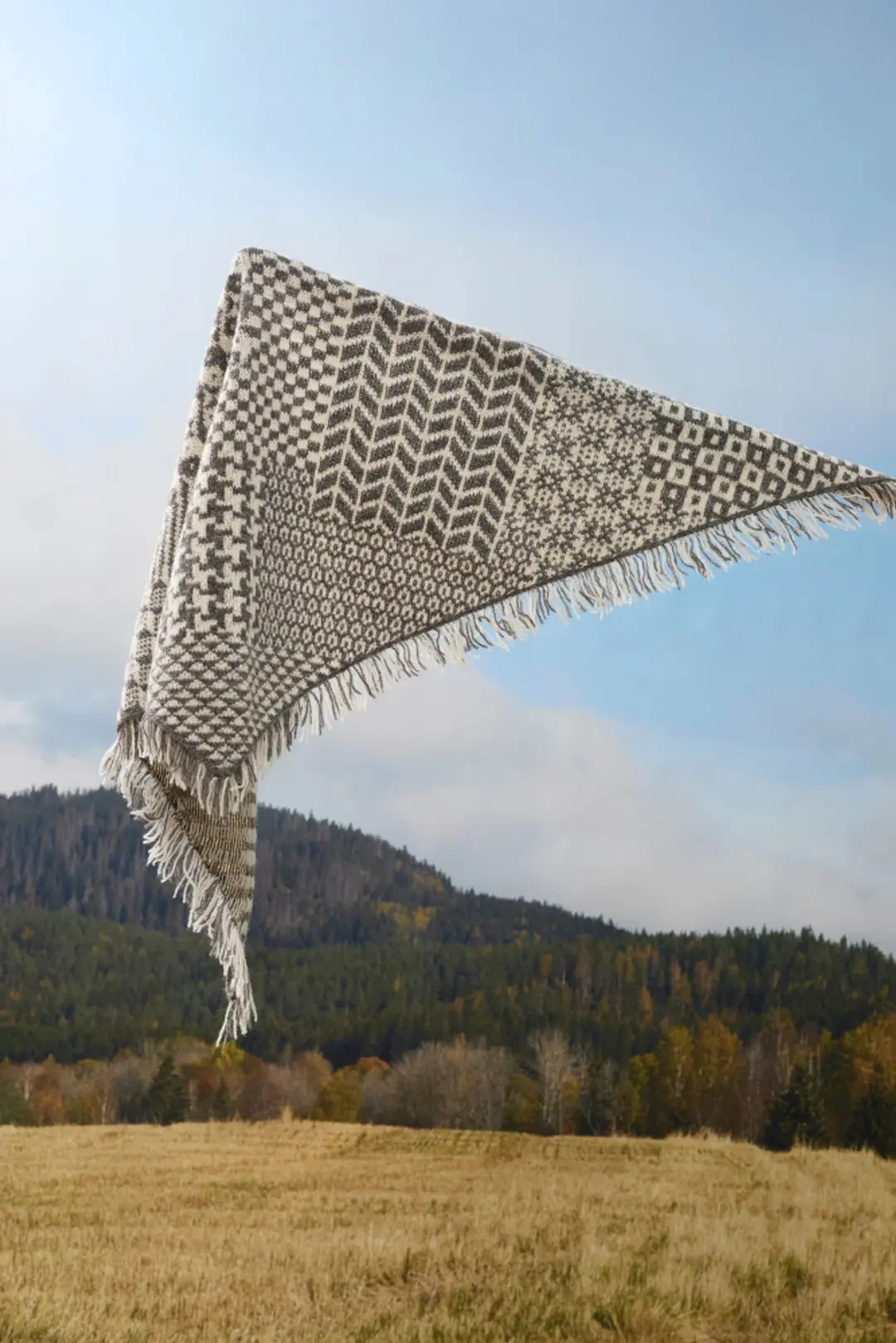 Et 455 Nordisk miks skjerf i grått og hvitt med frynser flyter uanstrengt mot et bakteppe av bølgende åser og klar himmel.