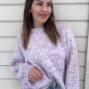 En kvinne med langt brunt hår, iført en lys lilla Cecilia XS-genser og blå jeans, står mot en hvit trevegg, ser litt til siden og smiler.