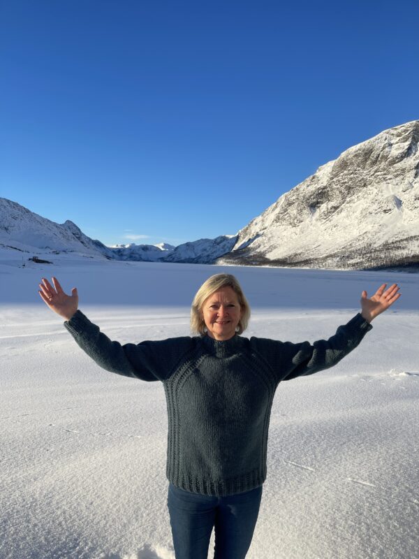 En person står med armene hevet på et snødekt landskap, omgitt av fjell under en klarblå himmel.