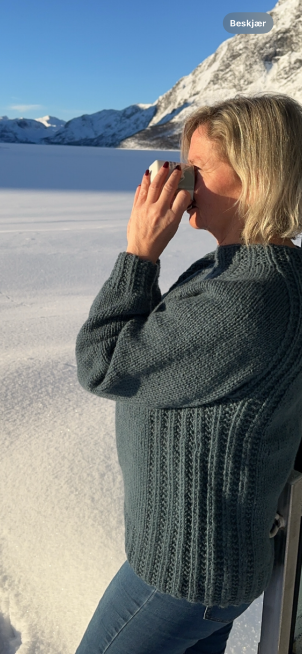 En person i en grå genser drikker av en kopp mens han står i et snølandskap med fjell i bakgrunnen.