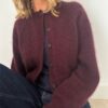 Iført en koselig burgunder Darcy cardigan over en mørk topp og jeans, sitter personen komfortabelt mot en vegg.