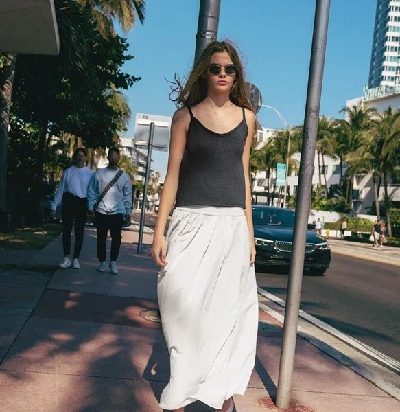 En kvinne i solbriller, iført 2504-04 Clementine-toppen og et hvitt skjørt, rusler langs et solfylt fortau omkranset av palmetrær og en imponerende bygning i bakgrunnen – hennes subtile eleganse minner om en morgenklementinsk glød.