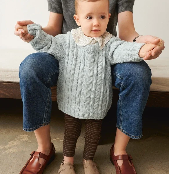 En baby i en lyseblå genser, iført brune leggings med 2505-02 Bunny Shorts og beige sko, står støttet av en voksen i en grå skjorte og blå jeans på sofaen.