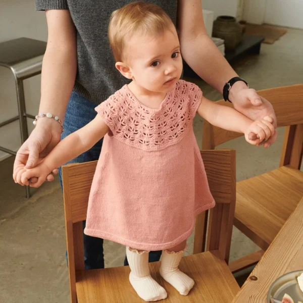 Et barn iført en 2505-02 Bunny shorts baby og en rosa kjole står på en stol med en voksens hjelp, ved siden av et bord dekket med tallerkener og mat.