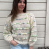 En person står mot en trevegg, iført en Bysse Genser i XL med fargerike aksenter på den hvite strikkegenseren og lyseblå jeans, en hånd tilfeldig i lommen.