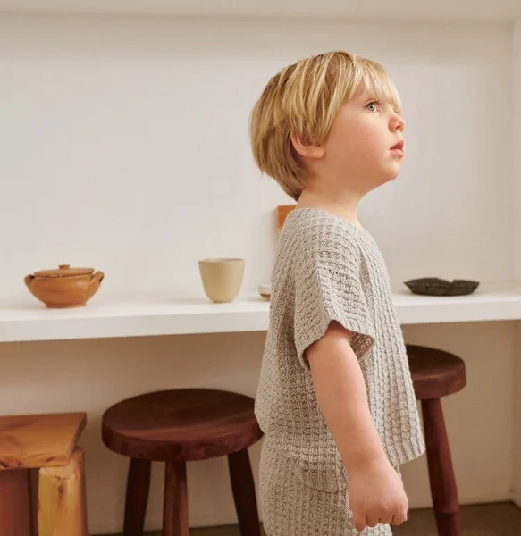 Et lite barn med blondt hår har på seg 2505-02 Bunny shorts baby i strikkede beige klær, stående sidelengs i et minimalistisk rom dekorert med hyller av keramikk.