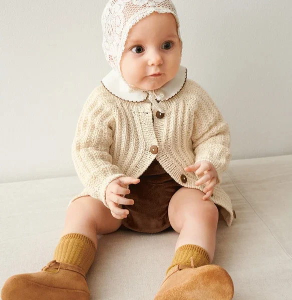 En baby i et søtt strikket antrekk og blondehette sitter på en lys overflate, iført en beige cardigan, 2505-02 Bunny shorts baby i brune, sennepssokker og solbrune sko.