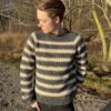 En mann iført Key Sweater Man står utendørs og ser sidelengs mot et bakteppe av nakne trær og sparsom vegetasjon.