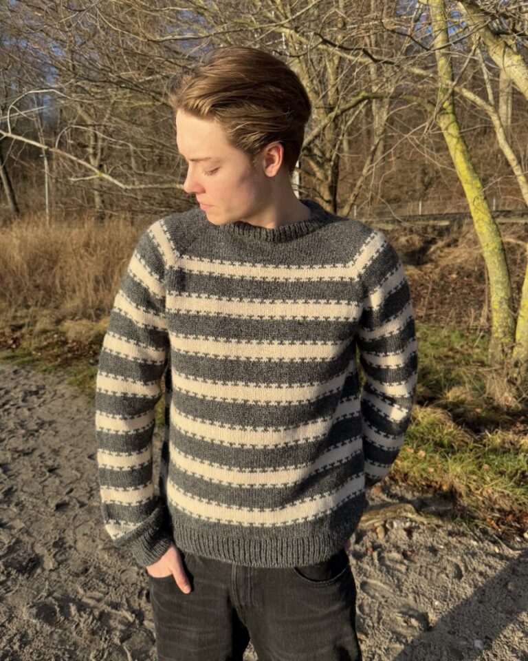 En mann iført Key Sweater Man står utendørs og ser sidelengs mot et bakteppe av nakne trær og sparsom vegetasjon.