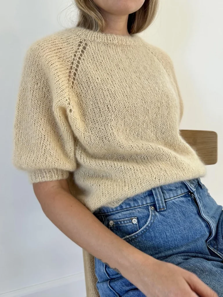 Sittende på en trestol mot en hvit bakgrunn har personen på seg "Puff" beige strikket kortermet genser sammen med blå jeans.