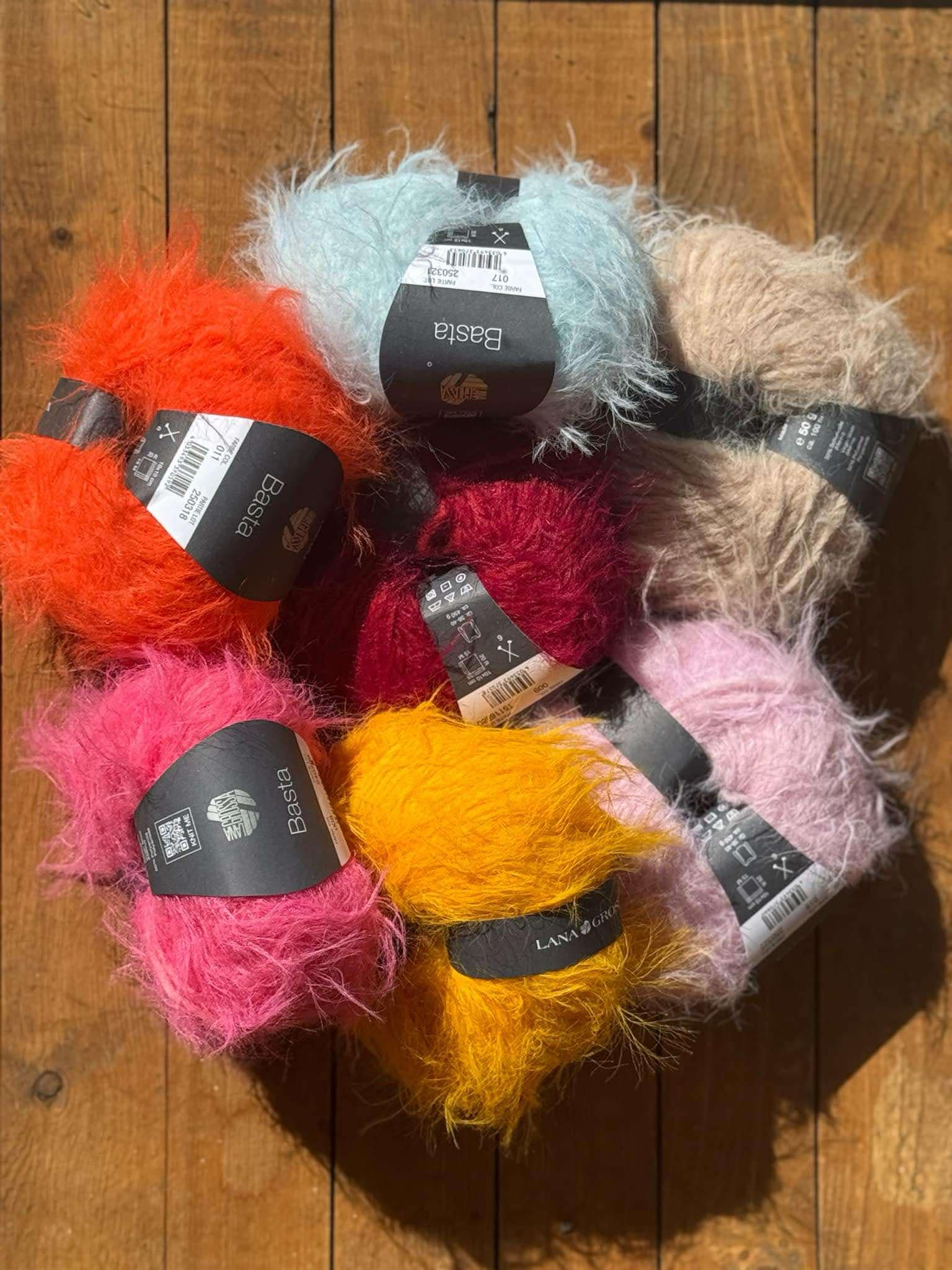 Syv nøster med fluffy rett garn i rødt, oransje, gult, rosa, lyseblått, burgunder og beige er arrangert på en treflate.