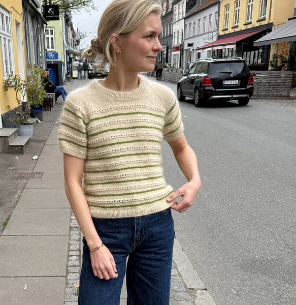 En kvinne står på et fortau iført Ida Tee – en kortermet, stripete strikkegenser – og jeans, med boligbygg og parkerte biler i bakgrunnen.
