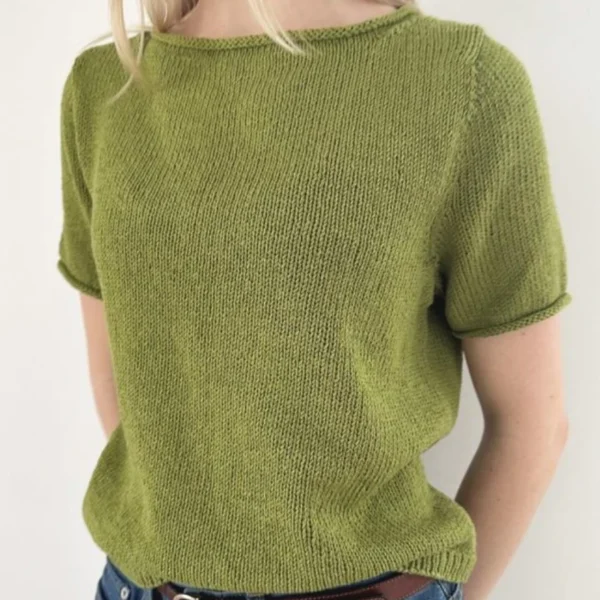 En person iført en kortermet grønn Vega Tee nedenfra strikketopp, blå jeans og et brunt belte står mot en enkel hvit bakgrunn.