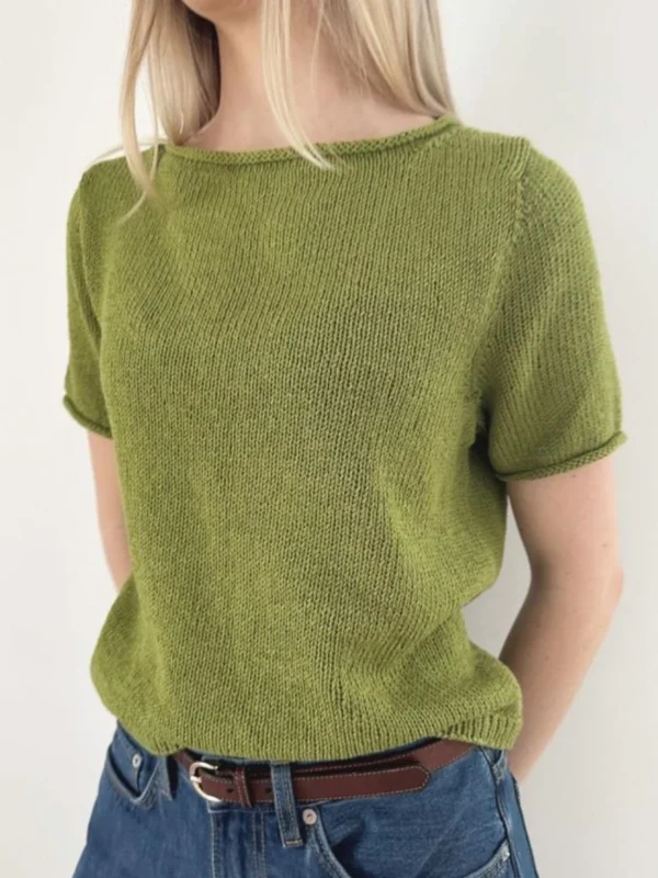 En person iført en kortermet grønn Vega Tee nedenfra strikketopp, blå jeans og et brunt belte står mot en enkel hvit bakgrunn.
