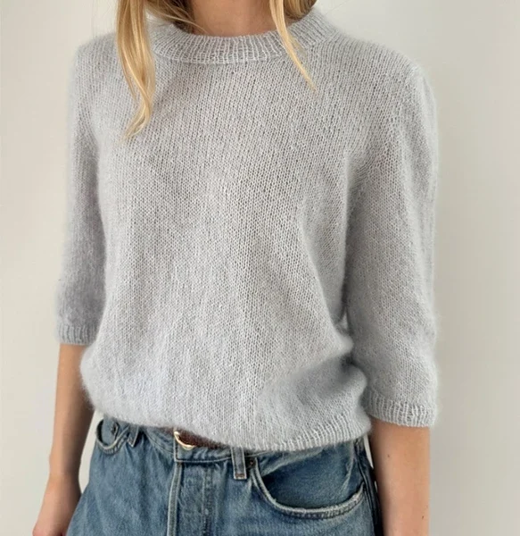 En kvinne i en lysegrå Maria-bluse med trekvartlange ermer og blå jeans med høy midje står mot en enkel, lys bakgrunn.