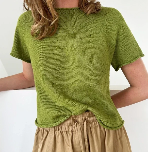 En person i en kortermet grønn Plain Yoke Tee 28 og et beige rynket skjørt står mot en ensfarget lys bakgrunn.