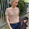 Utendørs holder en kvinne i mørke jeans en jakke og har på seg den kortermede beige Wave-t-skjorten, med grøntområder og et metallgjerde i bakgrunnen.