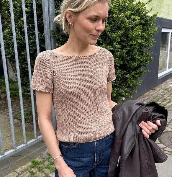 Utendørs holder en kvinne i mørke jeans en jakke og har på seg den kortermede beige Wave-t-skjorten, med grøntområder og et metallgjerde i bakgrunnen.