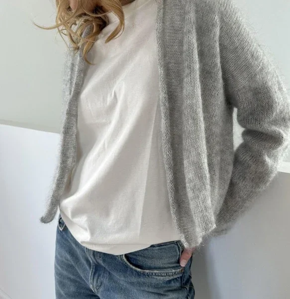Person iført hvit t-skjorte, lysegrå cardigan og blå jeans, står innendørs med én hånd i lommen.