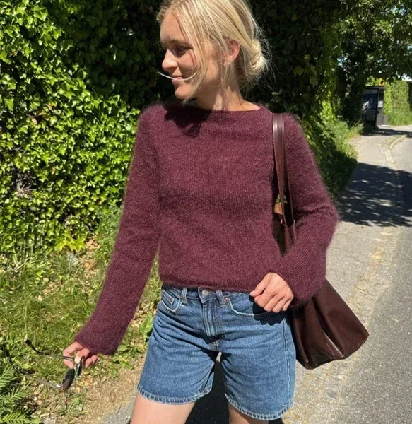 En kvinne iført en rødbrun genser og denimshorts står på et solrikt fortau med solbriller og en brun veske, med grøntområder og trær i bakgrunnen.